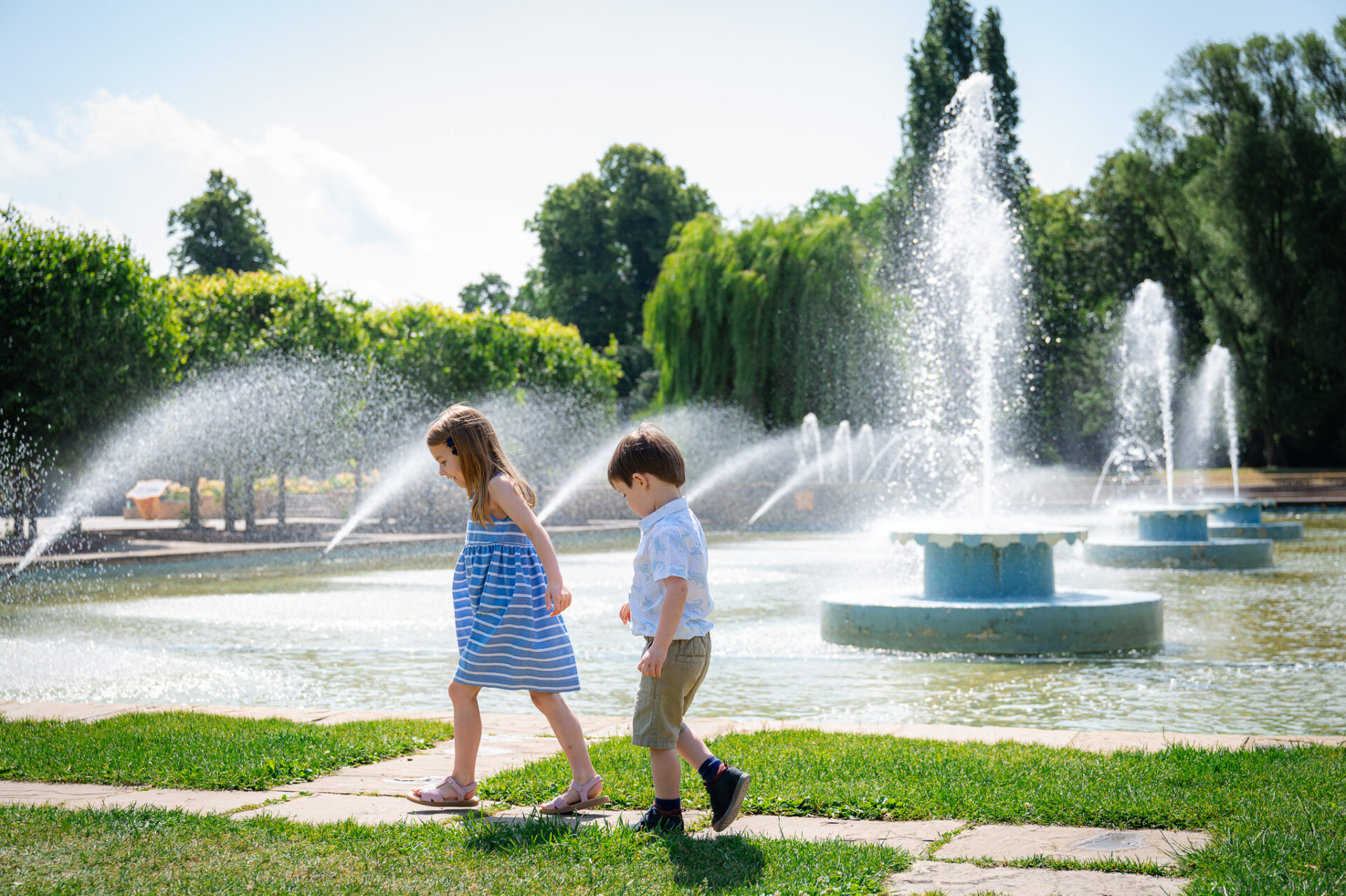 Children walk by a fountain on a sunny day, grassy park setting; girl in blue dress, boy in white shirt and shorts. Joyful, carefree atmosphere.