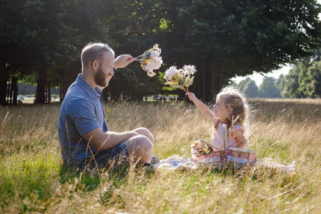 Little blonde girl holding flowers sitting on a picnic blanket with her father who is also holding flowers up high